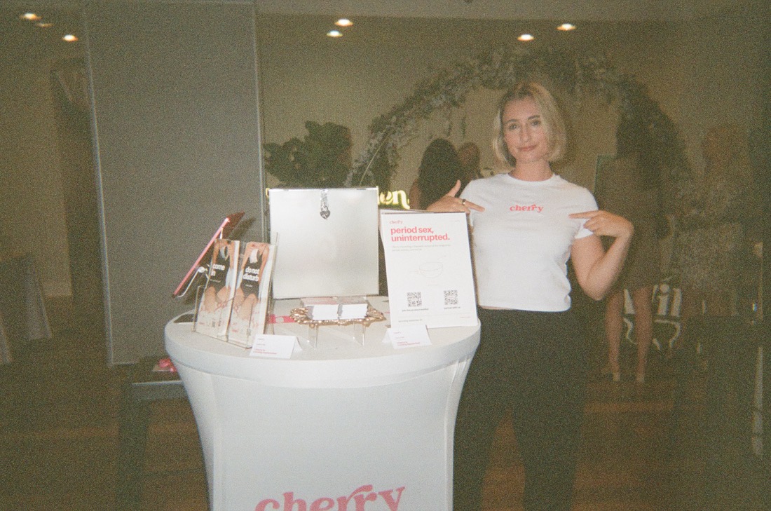 Anna in a cherry t-shirt pointing at the logo, beside a Cherry table with a sign that says period sex, uninterrupted