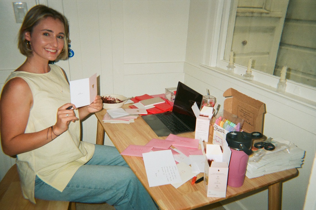 Anna at her kitchen table, smiling, holding a handwritten card, surrounded by pink envelopes, a laptop and boxes