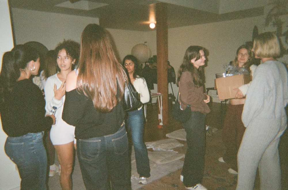 A room full of women standing and talking in small groups at a Cherry event, one of them carrying a cardboard box of supplies