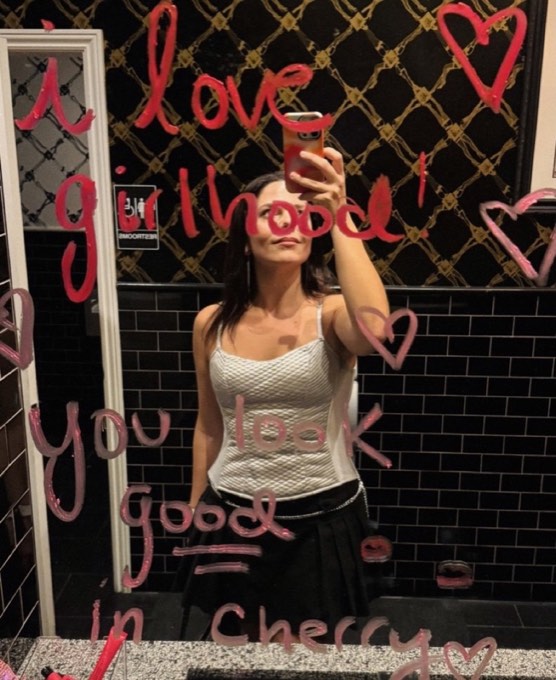 A woman taking a mirror selfie; the mirror is covered in lipstick writing that says i love girlhood and you look good in cherry