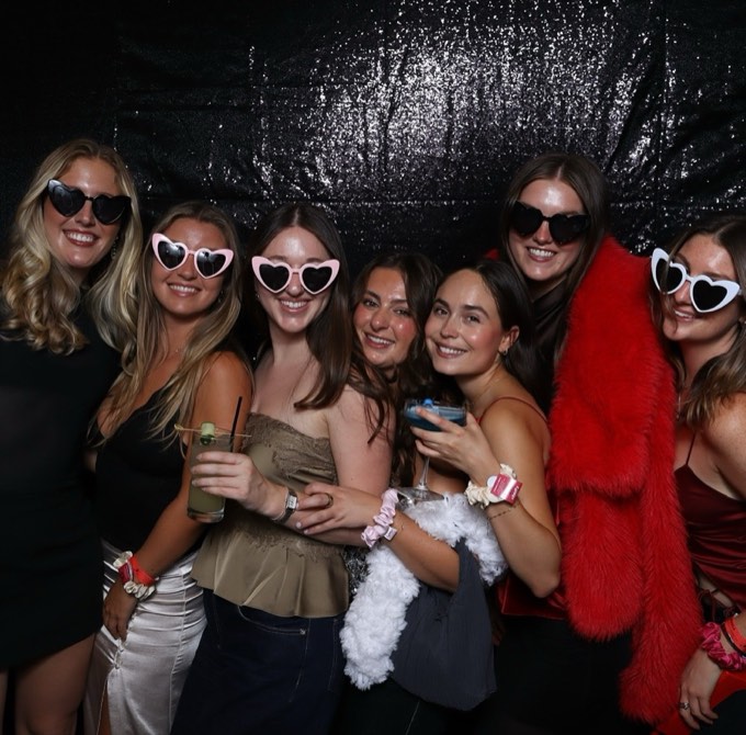 Seven women in the photo booth, most in heart-shaped sunglasses, one in a red faux fur coat