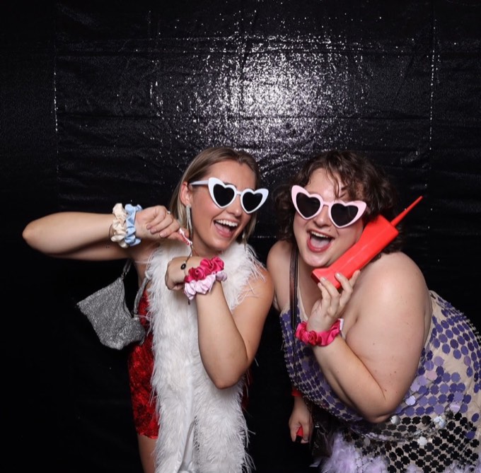 Two women in heart-shaped sunglasses laughing in the photo booth, one on a red prop phone
