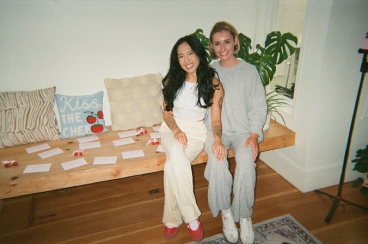 Two women smiling on a wooden bench next to a kiss the chef pillow and cherry clips